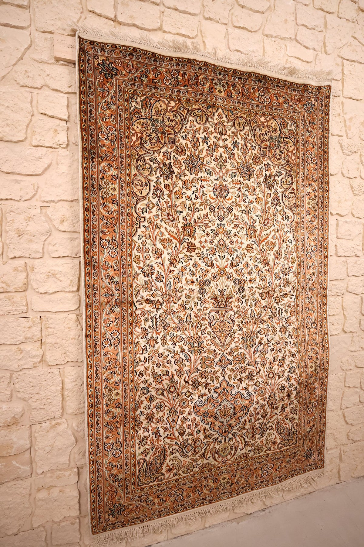 Tapis Cachemire beige en soie 159 × 95 cm, vue de biais sur le mur en pierre