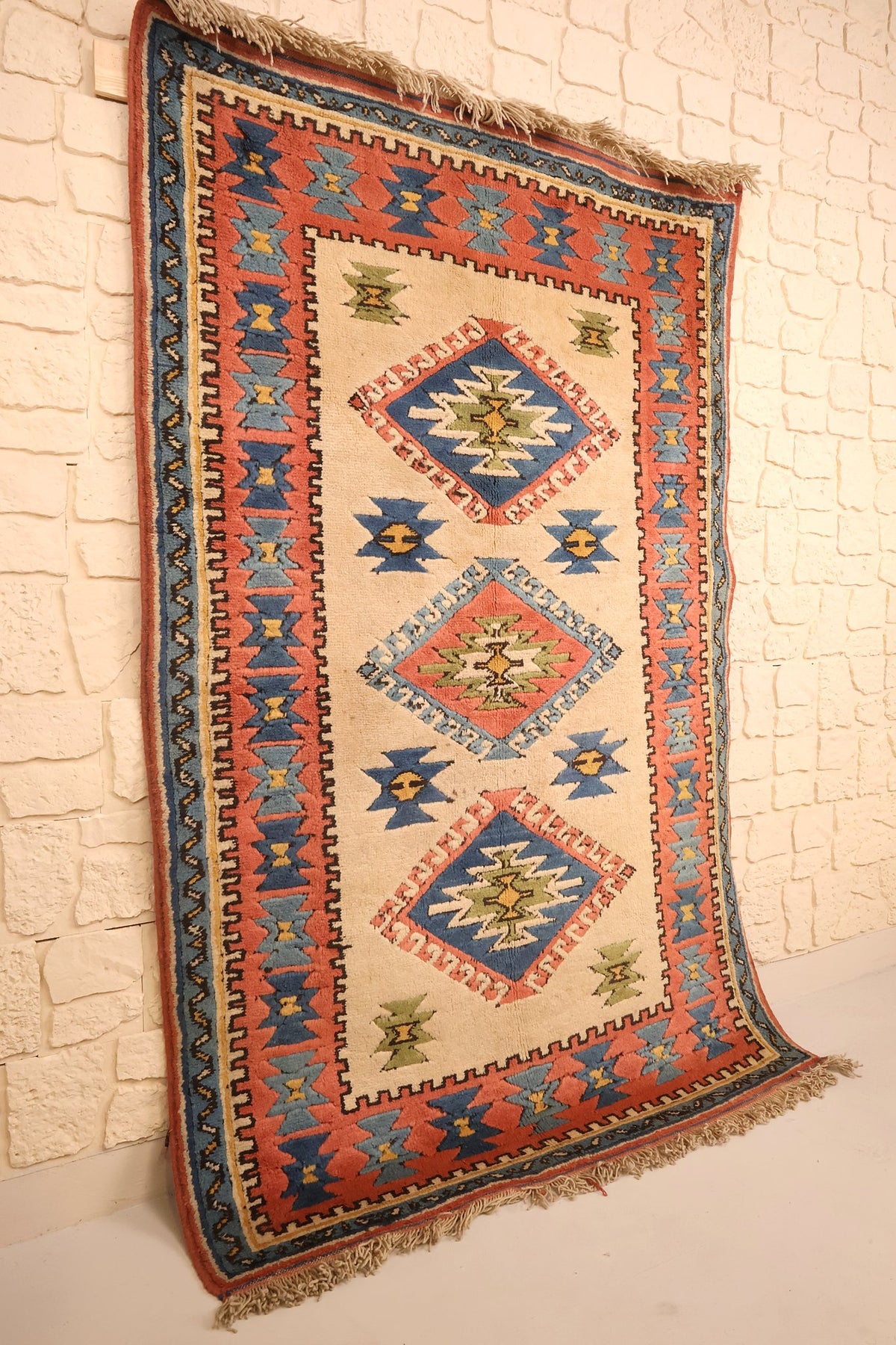 Tapis Konya rectangulaire beige et bleu, vue de biais contre un mur clair
